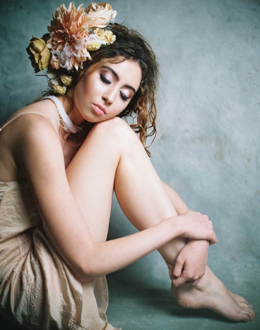 Female Model wearing floral headpiece resting her head on her legs while sitting on the floor.
