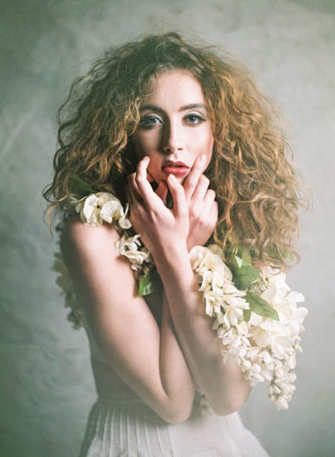 Female model looking at camera with arms crossed.  She is wearing a large floral necklace and has long curly hair.