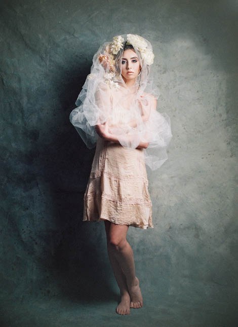 Female model standing and looking at camera wearing a long white veil and floral headpiece.