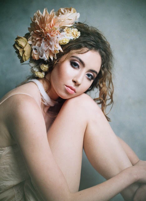 Female model with blue eyes wearing a floral headpiece sitting on the ground and resting her head on her legs.