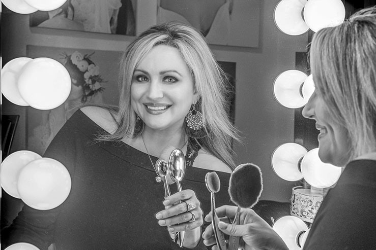 Ashlee Rice holding artis makeup brushes and smiling into a mirror in black and white.