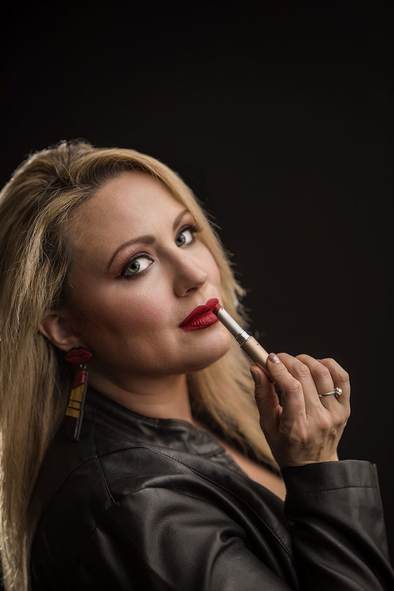 Ashlee Rice looking at the camera provocatively while putting on dark red lipstick in front of a black background.