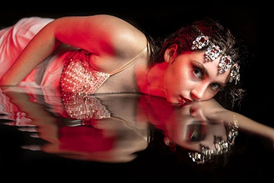 Girl laying in water wearing a dress and tiara,  Half her face is in the in the water and half is looking at the camera.